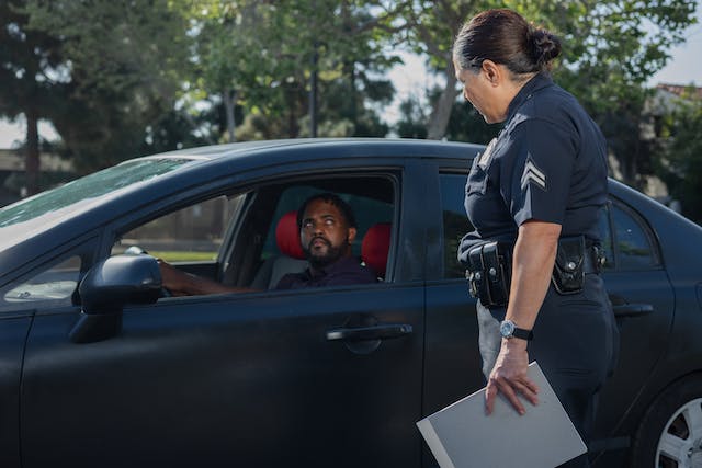 A police officer stands beside a black car, discussing Texas insurance laws with a man in the driver's seat. The sunny day and trees in the background frame their exchange. Clipboard in hand, the officer ensures the driver understands that driving without insurance is illegal in Texas.