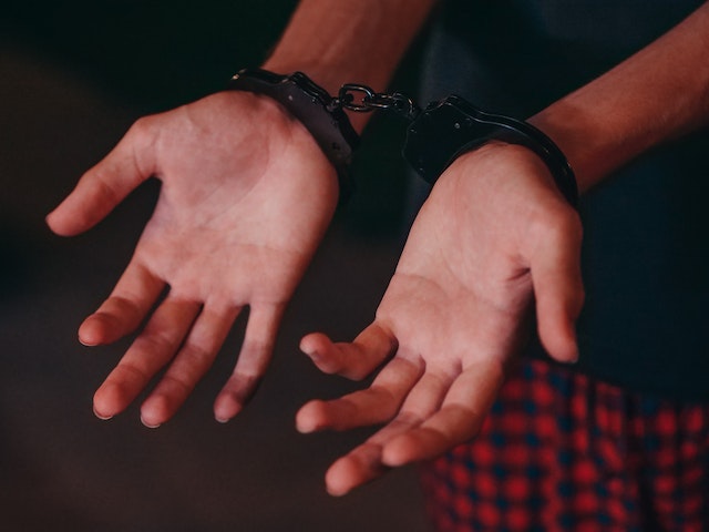 Close-up of a person's hands with palms facing up, wearing handcuffs—a stark symbol often seen amid the rising tide of crime in Texas. The person dons red and black checkered pants, set against a blurred, dark background.
