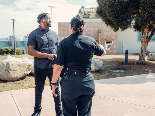 A man and a woman in uniform stand facing each other outdoors near a building. The woman gestures with her hand as if instructing on a field sobriety test, and they appear to be engaged in conversation.
