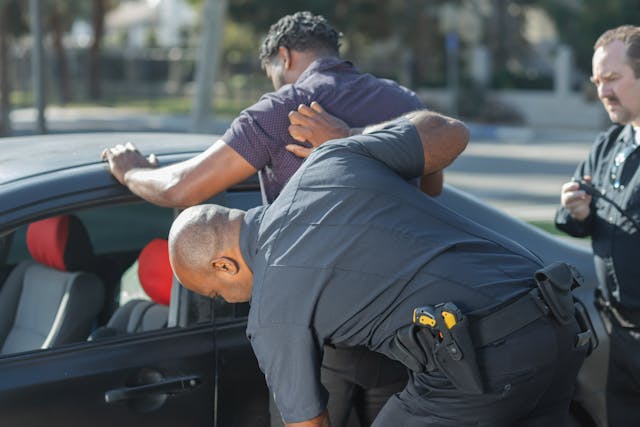 A police officer conducts a vehicle search on a man leaning against a car, while another officer stands nearby. The scene unfolds outdoors on a sunny day, capturing the essence of routine law enforcement duties.
