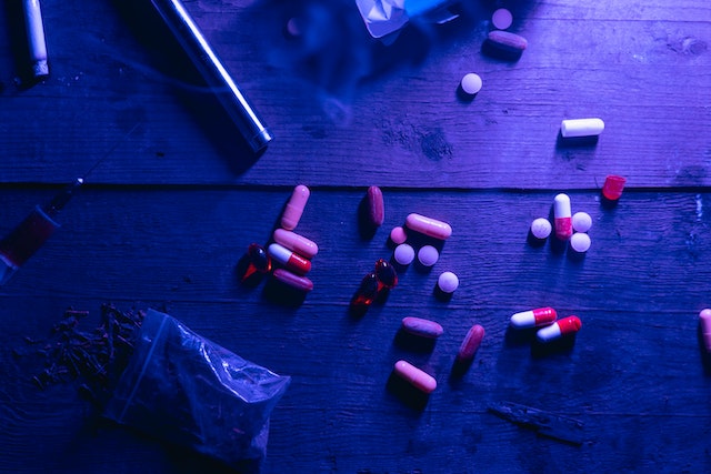 A dimly lit wooden surface displays scattered pills, capsules, a syringe, and a small bag containing a substance—an unsettling scene of constructive possession. The moody atmosphere is heightened by purple lighting casting ominous shadows.