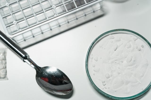 Close-up of a spoon next to a Petri dish containing a white powdery substance, reminiscent of the strict Texas cocaine laws, with a white grid structure partially visible in the background on a white surface.