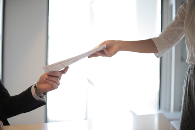 In a brightly lit office setting, a convicted felon hands over a document to another person across the table. The recipient's arm is partially visible, creating an intriguing snapshot of transition and trust in unexpected places.