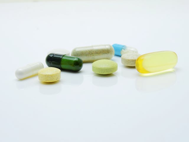 A variety of pills and capsules, resembling controlled substances in their diverse colors and sizes, are scattered on a smooth white surface. This assortment features yellow, green, and blue capsules alongside round tablets.