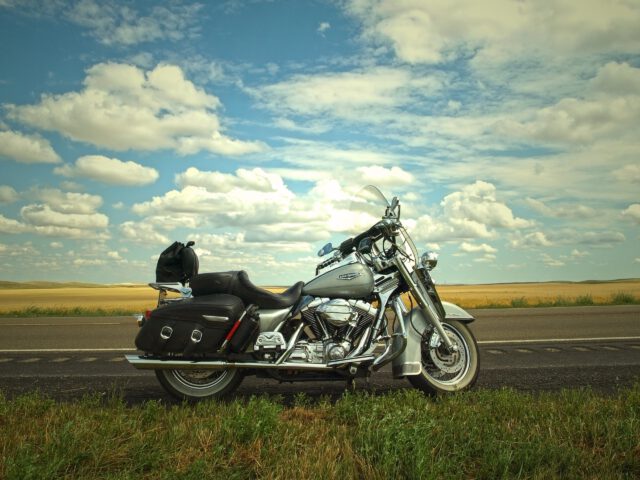 A silver and black motorcycle parked on the side of a Texas road, with vast open fields and a cloudy blue sky in the background. The saddlebags are visible, offering motorcyclists a glimpse into this expansive and serene landscape.