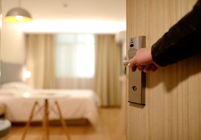 A person opens a door to a hotel room in Texas. The room features a neatly made bed, soft lighting, and curtains on the far wall, creating a welcoming atmosphere despite recent hotel accidents that have alerted guests to be cautious.