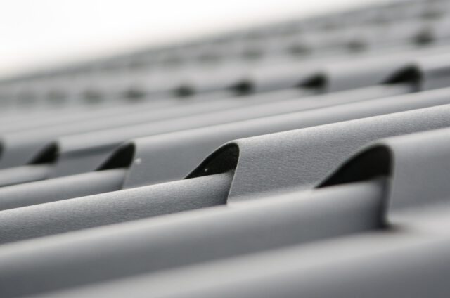 Close-up of a grey, corrugated metal roof showing the parallel ridges and grooves extending into the distance, creating a pattern of lines and shadows. The focus is on the texture and structure, highlighting its resilience against unexpected damages.