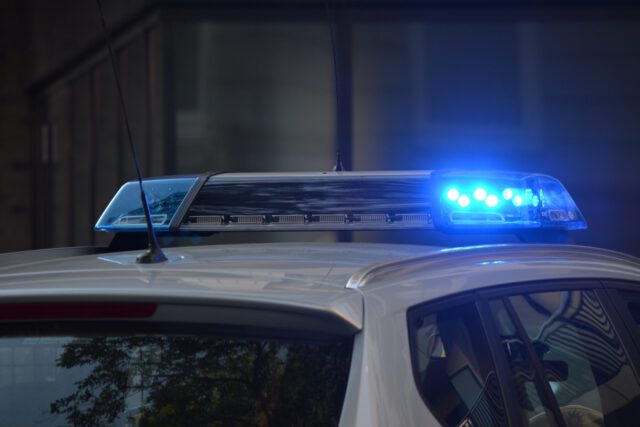 A police car with blue emergency lights flashing on top is parked in a dim urban setting, responding to an armed robbery. The lights illuminate the surrounding area, reflecting off the car's roof, casting an urgent glow on the unfolding scene of crime.
