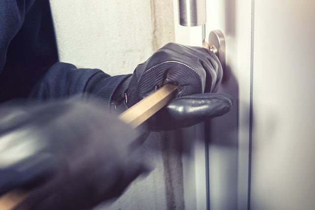 A person wearing black gloves uses a crowbar to pry open a door lock, clearly depicting a burglary in progress. This scene hints at the seriousness of such felonious activities commonly reported in Texas neighborhoods.