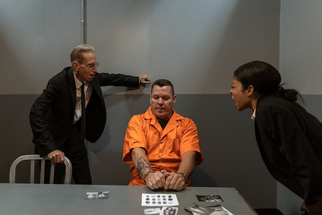 In a dimly lit Texas interrogation room, two detectives confront a man in an orange prison jumpsuit. One detective leans over the table, asserting authority, while the other sits across, both appearing confrontational. The tension is palpable amid photographs on the table as they discuss his rights.