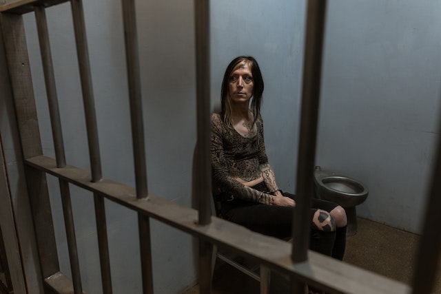 A person with long dark hair sits on a chair in a dimly lit Texas jail cell, visible through metal bars. The concrete walls and a small metal toilet highlight the austere environment. The person is wearing a patterned top and has a somber expression, contemplating their need for a bail bond.