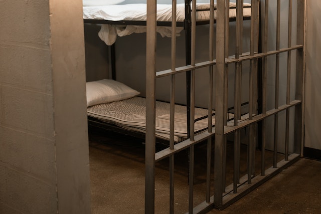 A dimly lit prison cell with metal bars and a concrete floor awaits those caught violating parole terms. Inside, two bunk beds with striped mattresses and white pillows sit against the plain walls, enhancing the stark atmosphere of consequences after a Texas parole misdemeanour.