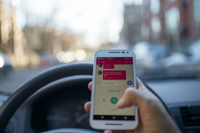 A person holds a smartphone with a messaging app open while sitting in the driver's seat of a car, capturing the risky act of texting while driving. The steering wheel and a street view through the windshield in Texas are visible, with blurred trees and vehicles in the background.