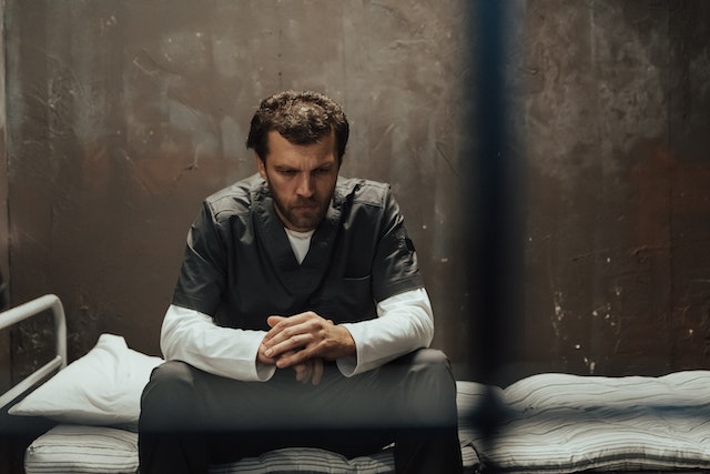 A man in scrubs sits on a bed in a dimly lit Texas room with his hands clasped, appearing contemplative. The bare, textured walls and metal bed frame echo the weight of being falsely accused of domestic violence.