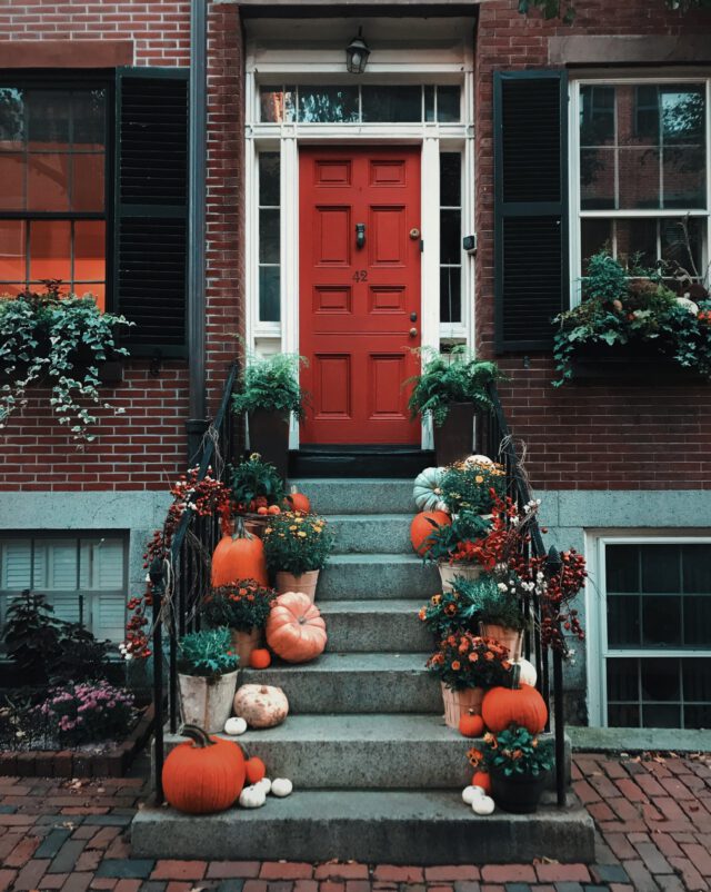 A brick townhouse with a red door, flanked by black shutters, invites Texas homeowners. The steps are adorned with pumpkins, gourds, and fall flowers in pots, crafting a festive autumn scene while ensuring Halloween safety tips are considered for a secure celebration.