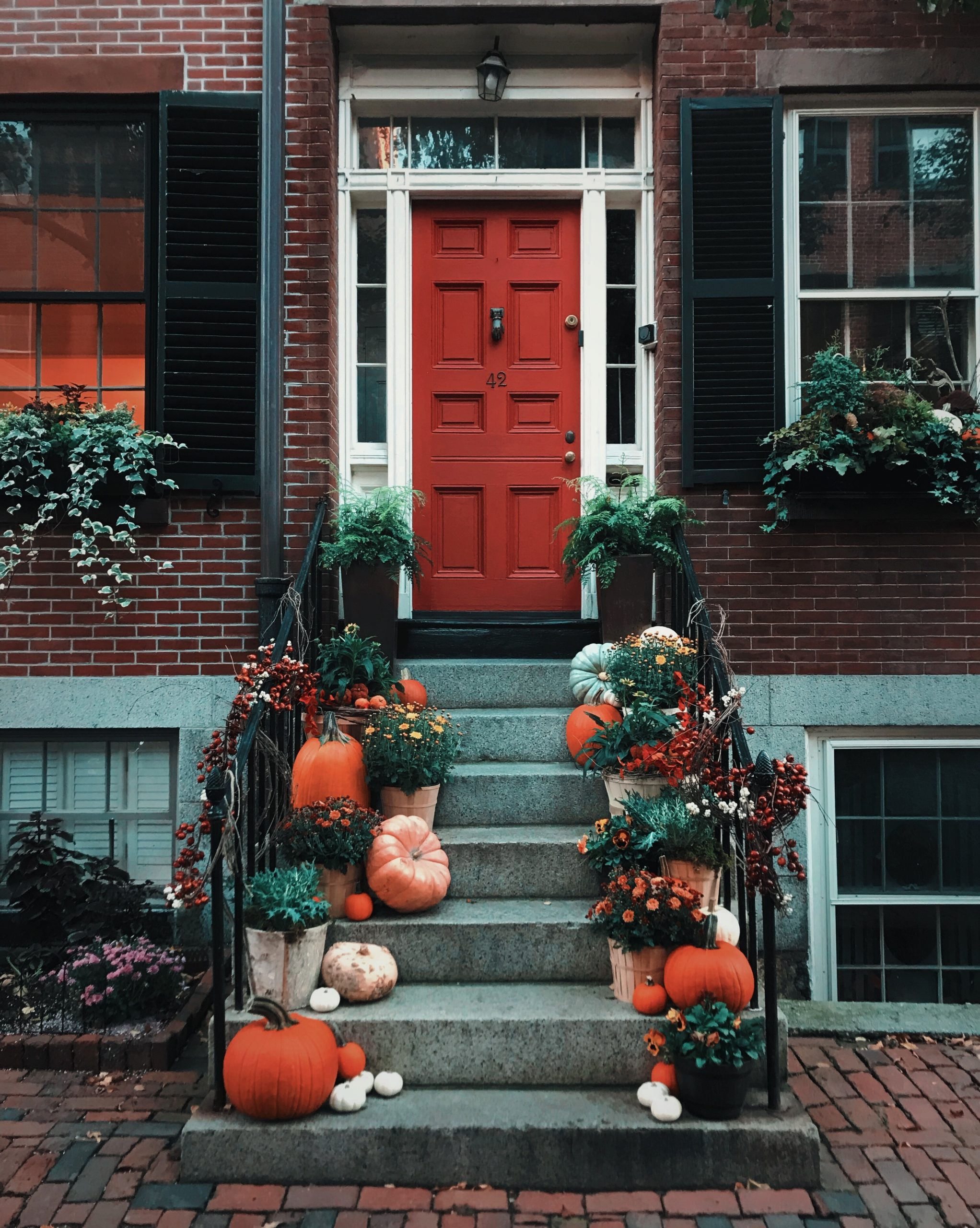 A brick townhouse with a red door, flanked by black shutters, invites Texas homeowners. The steps are adorned with pumpkins, gourds, and fall flowers in pots, crafting a festive autumn scene while ensuring Halloween safety tips are considered for a secure celebration.