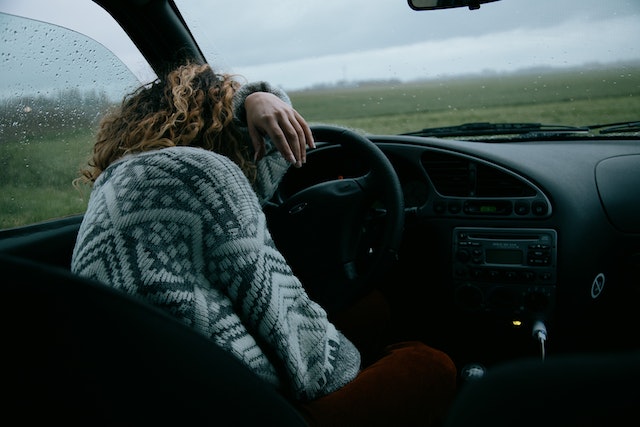 A person with curly hair wearing a patterned sweater is resting their head on the steering wheel inside a car in Texas. It's raining outside, and the windows are covered in rain droplets, giving the impression they might be sleeping or perhaps reflecting deeply while avoiding a DWI incident.