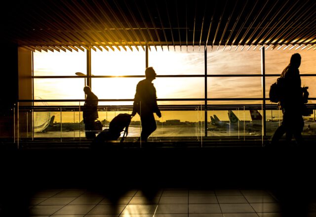 Silhouettes of travelers with luggage navigate through a bustling Texas airport terminal. The large windows backdrop a vibrant sunset, casting shadows on the floor, while airplanes await on the tarmac.