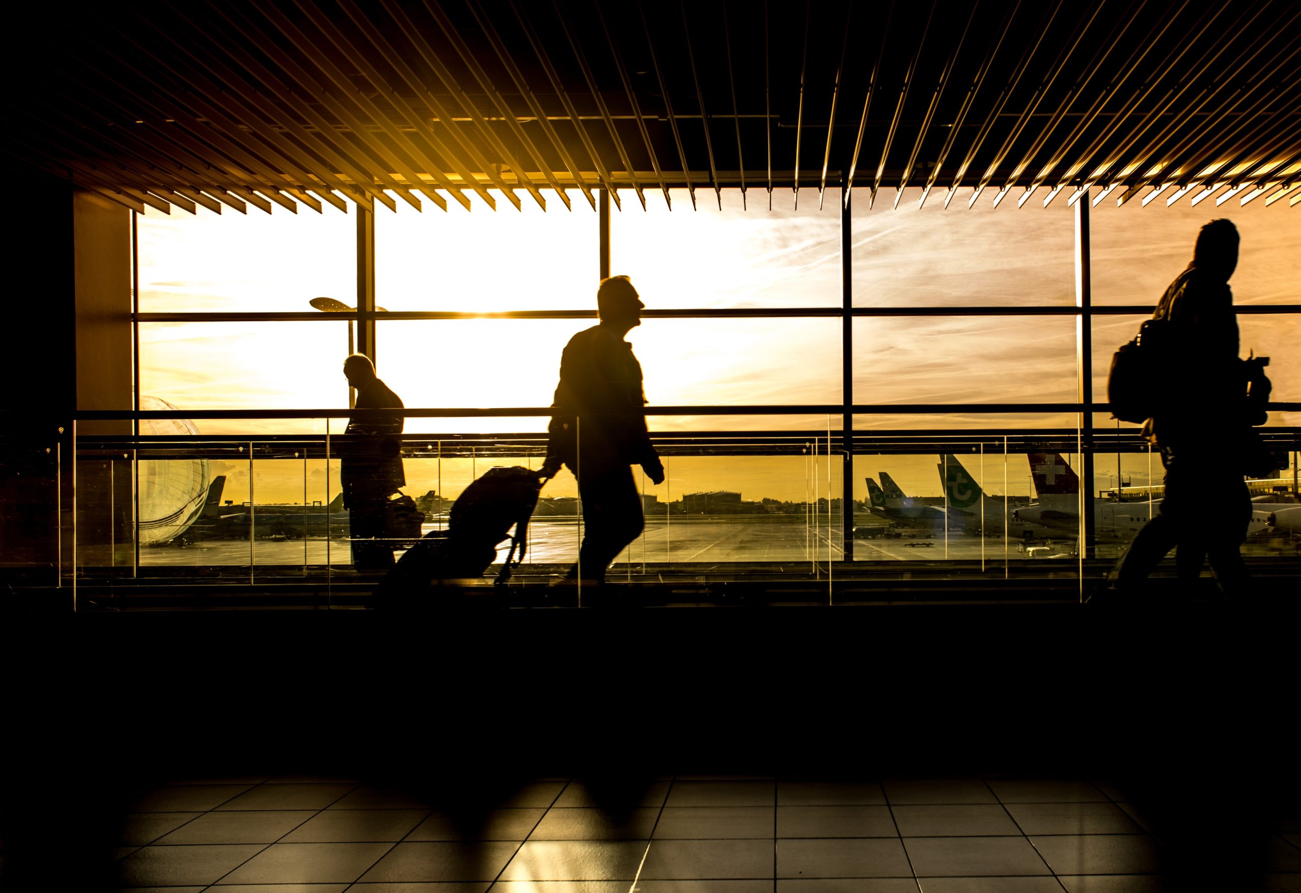 Silhouettes of travelers with luggage navigate through a bustling Texas airport terminal. The large windows backdrop a vibrant sunset, casting shadows on the floor, while airplanes await on the tarmac.