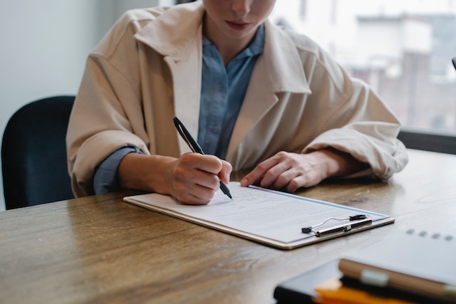 A person in a light coat sits at a wooden desk, writing on a clipboard with a pen. The clipboard holds several sheets of paper detailing the career impact of a Texas felony conviction. The background shows a blurred view of a window and buildings.