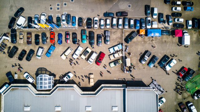 Aerial view of a bustling Texas parking lot with numerous vehicles parked in rows. Small groups of people are walking around, and a few colorful tents are set up on the right side of the image, showcasing what you should know about lively outdoor events beside a gray building.
