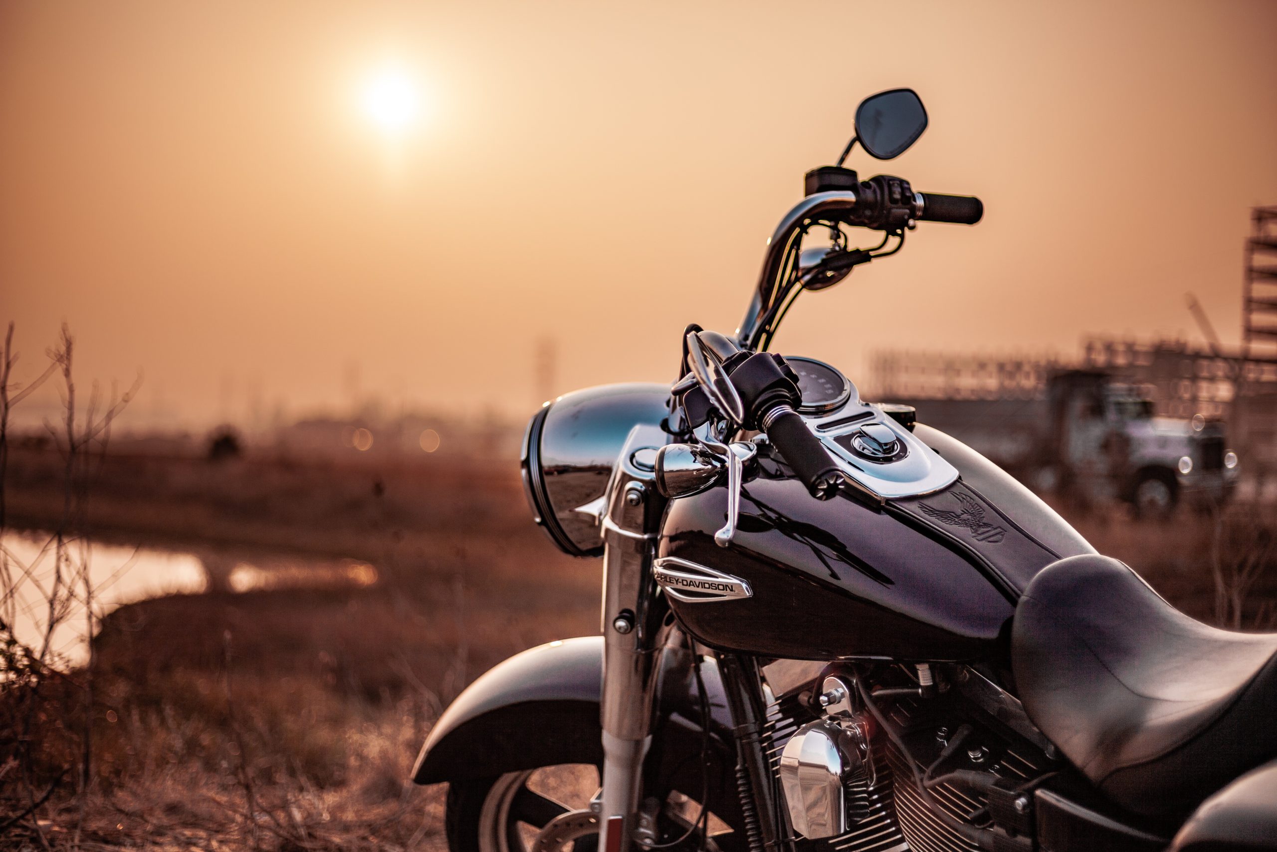 A black motorcycle, a reminder of post-accident actions, rests on a grassy area as the sun sets. The sky glows orange beyond a distant truck, with grass and a small body of water nearby.