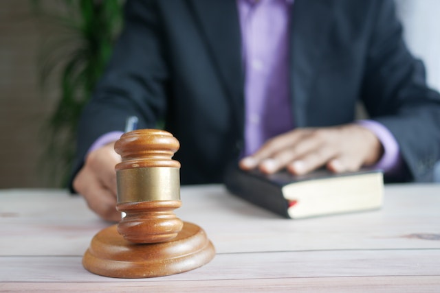A person in a suit holds a book with one hand while reaching for a wooden gavel on the table, embodying the legal term 