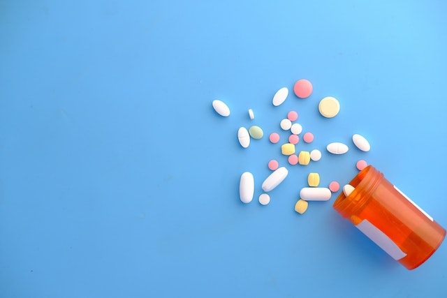 A variety of colorful pills and tablets, representing critical prescription medication, spill out of an orange bottle onto a blue surface.