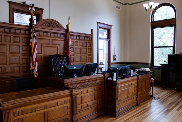 A courtroom with wooden paneling and furniture exudes a sense of gravitas. The empty judge's bench, awaiting the next case, is equipped with computer monitors and framed by an American flag. Large arched windows let in natural light, enhancing the room's well-lit classic design.