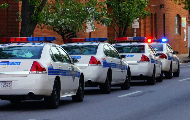 Four police cars with flashing lights are parked in a row along a tree-lined street, perhaps due to a probation violation. A brick building looms in the background, setting the scene for potential Texas consequences.