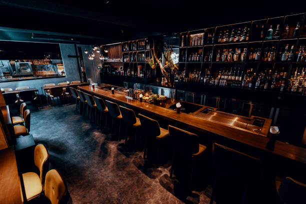 A dimly lit, modern bar in Texas features high chairs lining a long wooden counter. Shelves stocked with bottles gleam under soft lighting, crafting a cozy atmosphere. Tables are arranged in the foreground while a large mirror reflects the scene—despite whispers of a poor lighting accident lawsuit.