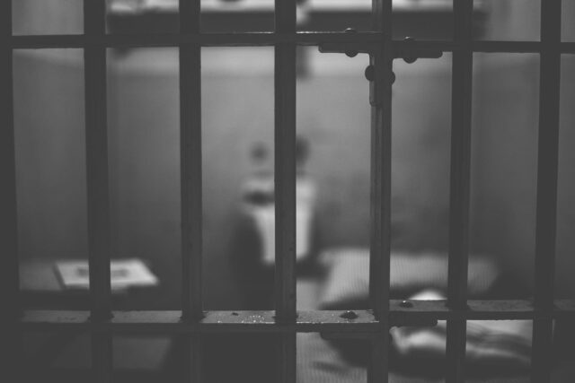 Black and white image of a Texas prison cell, with focus on the bars in the foreground. Inside, a blurred toilet and bed are visible, conveying the sparseness of the interior often found in facilities housing those convicted of manslaughter.