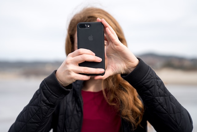 In Texas, a person with long hair, wearing a black jacket and burgundy shirt, holds a black smartphone up to record police interactions, shielding their face in an outdoor setting.