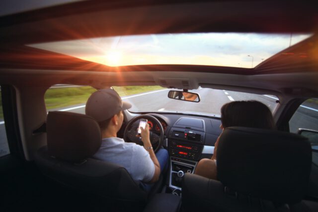 A person wearing a gray cap drives a car on an open road at sunset, careful to avoid the pitfalls of texting while driving. Their companion sits beside them, enjoying sunlight streaming in through the sunroof, casting a warm glow over the interior.