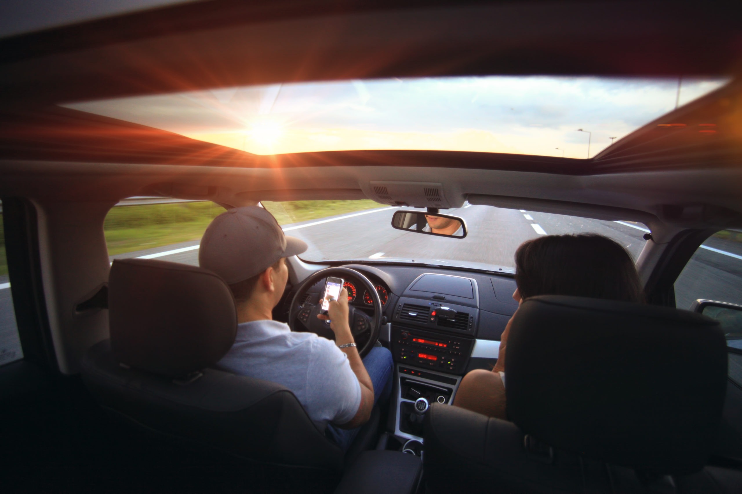 A person wearing a gray cap drives a car on an open road at sunset, careful to avoid the pitfalls of texting while driving. Their companion sits beside them, enjoying sunlight streaming in through the sunroof, casting a warm glow over the interior.