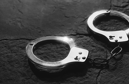 Close-up of a pair of handcuffs lying on a dark, textured surface. The handcuffs are metallic, reflecting light on their surface, casting subtle shadows on the background.