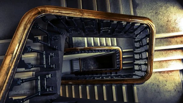 A top-down view of a spiral staircase in Texas features dark railings and wooden handrails. The worn steps, combined with dramatic lighting, create shadows that evoke depth and mystery. This scene highlights the stairwell's history, whispering tales of past incidents and what to know about its safety.