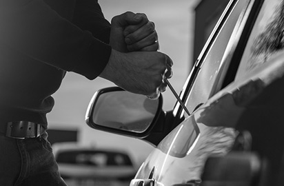 A person attempting to break into a car using a tool, focusing on the hands and the side mirror. The image is in black and white.