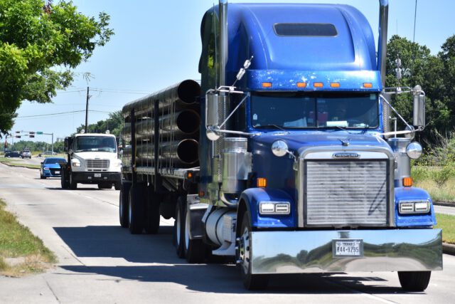 A blue semi-truck, expertly navigated by seasoned truck drivers, hauls large pipes down a sunny road. Another truck and a car follow closely behind. Green trees line the left side of the street, and a traffic light is visible in the distance.