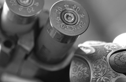 A close-up, black-and-white image of two shotgun shells loaded into the twin barrels of an ornate firearm. The shotgun features intricate engraved detailing on the metal, adding an elegant design touch.