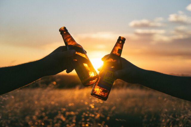 Two silhouetted hands clink beer bottles against a vibrant Texas sunset. The sun's golden light creates a warm glow through the bottles, set in an open field with a clear sky, reminding all to celebrate responsibly amidst ever-important DUI laws.