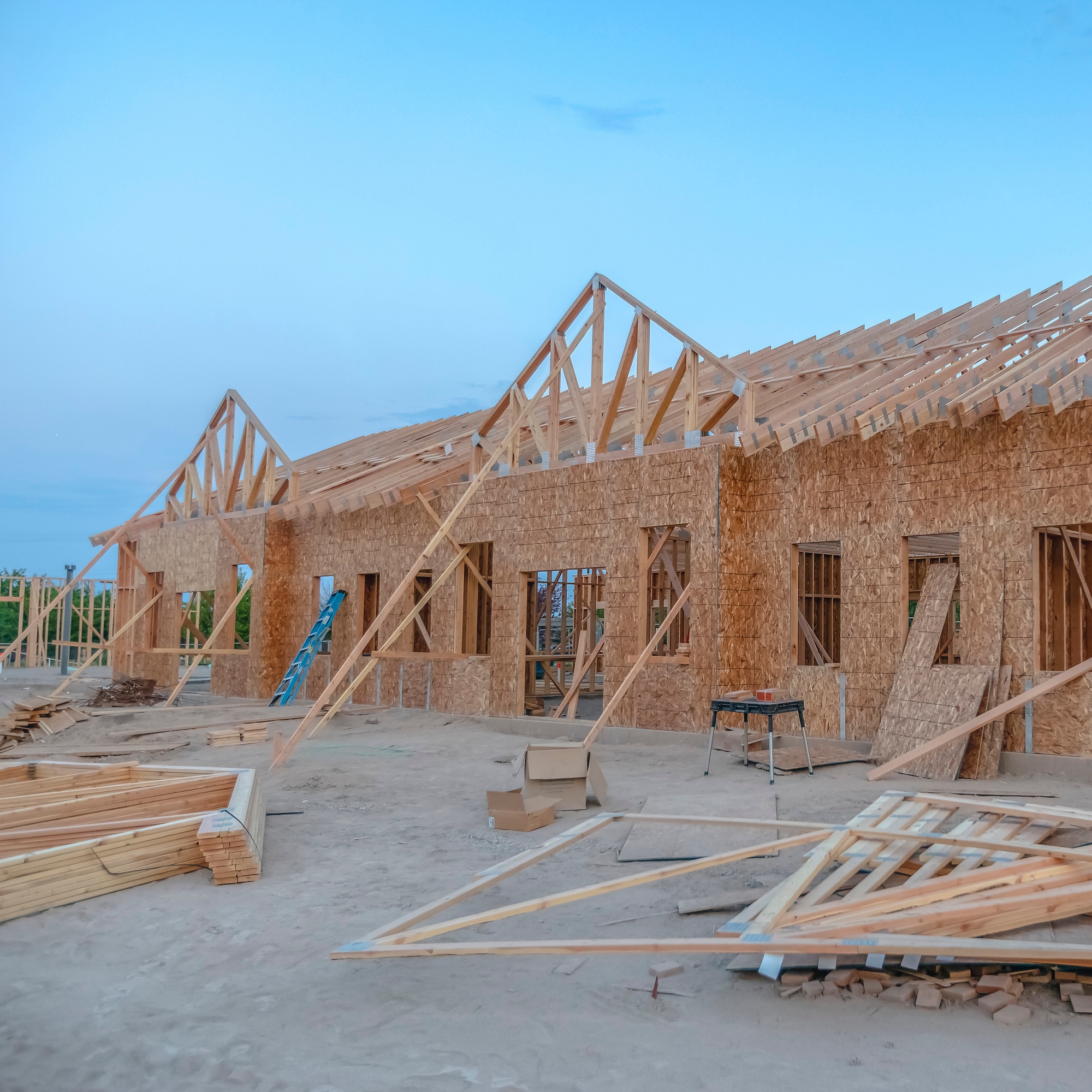 A partially constructed wooden house stands on a sandy lot. The frame is complete with trusses visible against a clear blue sky. Construction materials, including planks and beams, are scattered around the site.