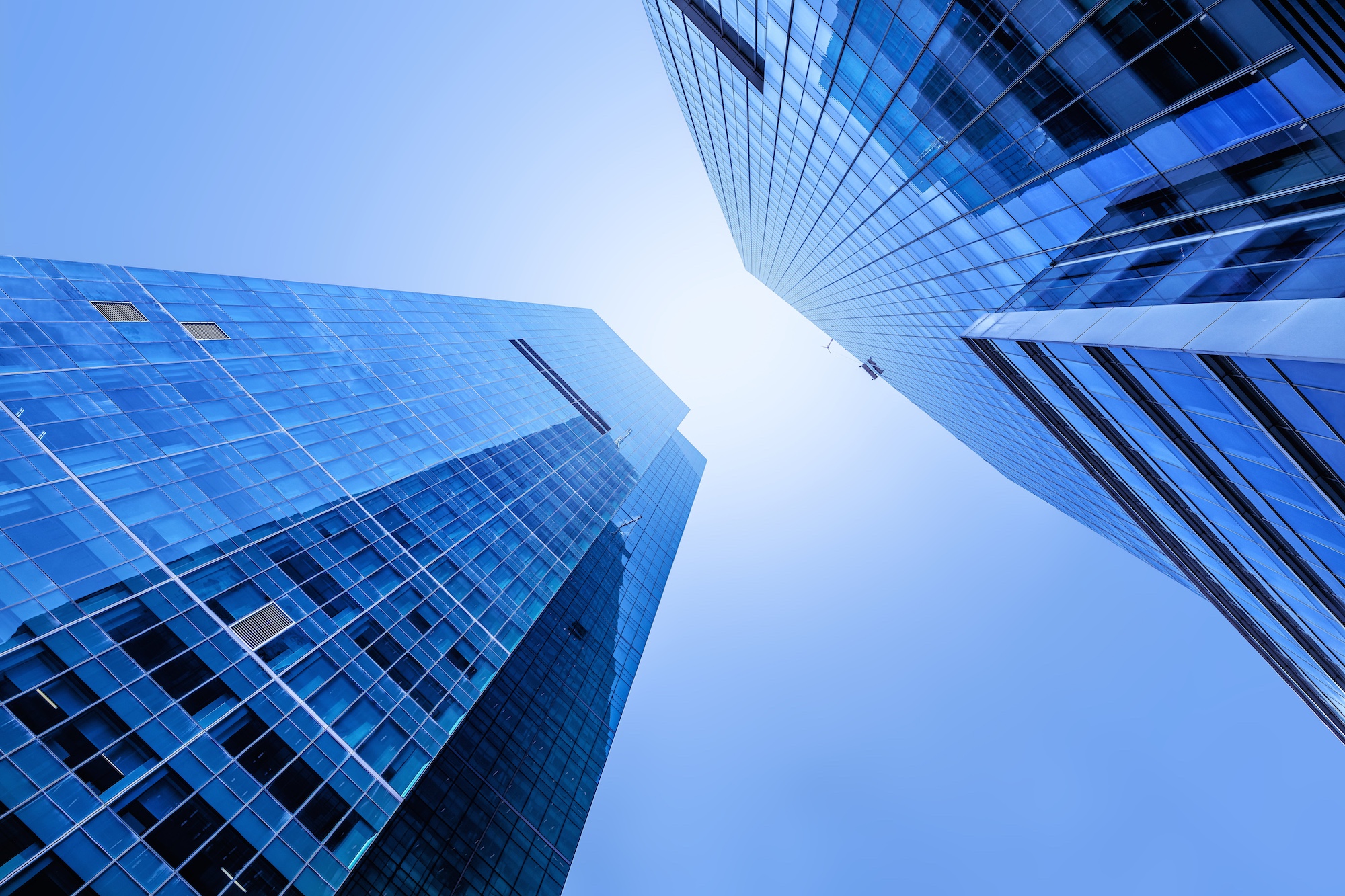 A low-angle view of two tall, modern skyscrapers with glass facades reflecting the sky. The buildings converge towards the center, creating a sense of height and perspective against a clear blue sky illuminated by sunlight.