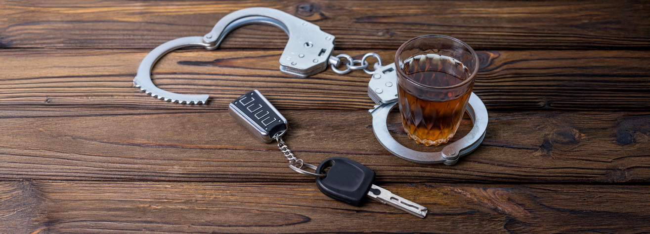 A pair of handcuffs is fastened around a glass of brown liquid, resembling alcohol, resting on a wooden surface. A car key and keychain are also connected to the handcuffs, symbolizing drink driving consequences.