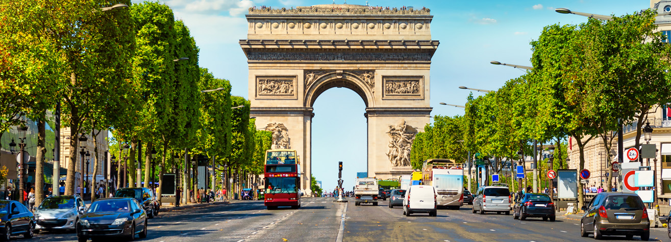 A view of the Arc de Triomphe in Paris, France. The wide avenue, lined with green trees, is bustling with traffic, including cars and a red double-decker bus. The sky is clear and blue.