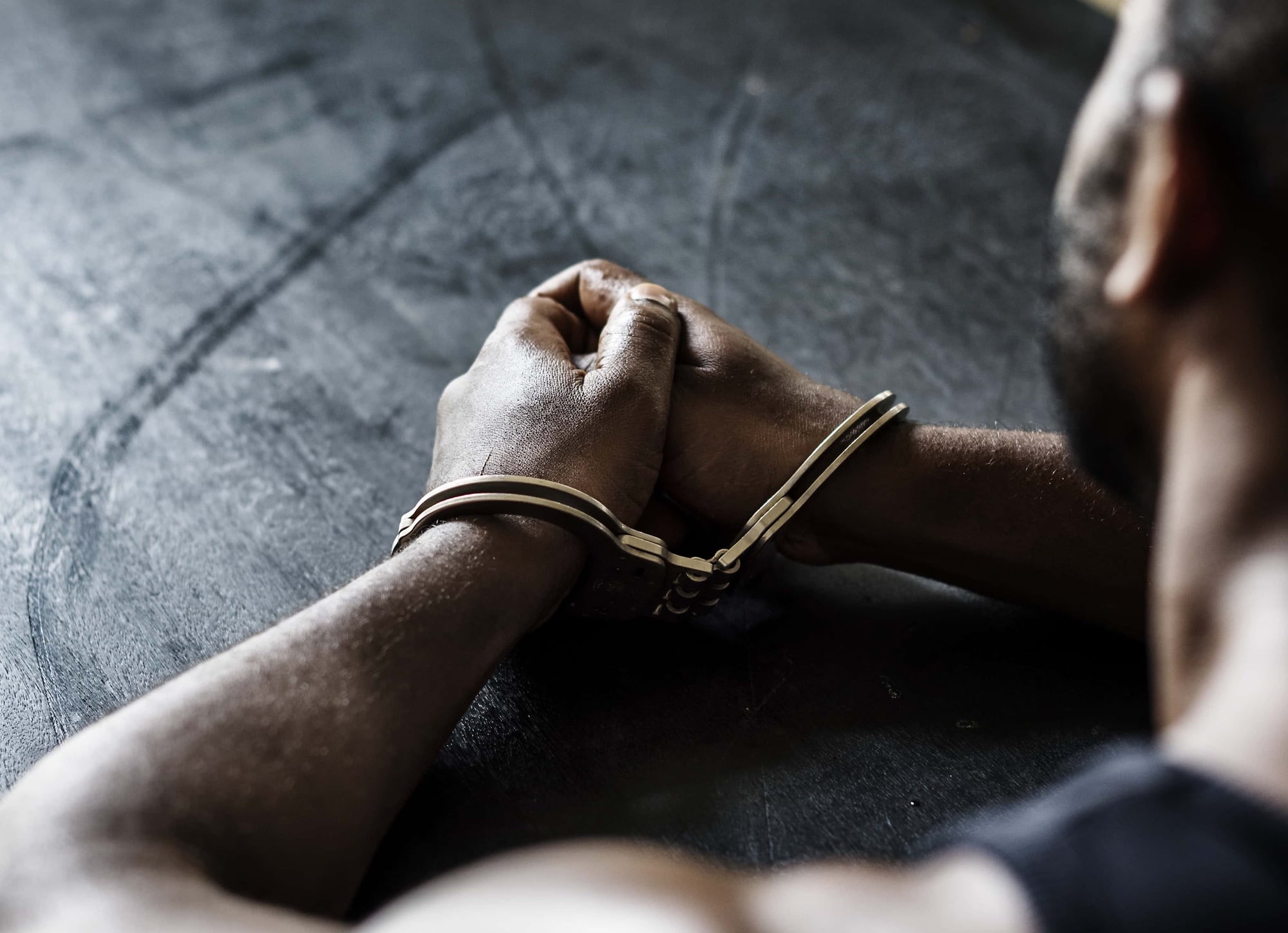 A person with dark skin sits with arms resting on a dark surface, hands clasped together and secured with zip ties. The focus is on the hands and arms, depicting restraint and tension. The background is blurred.