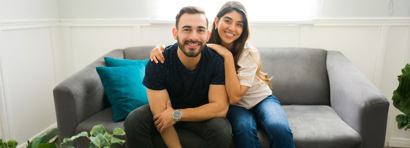 A smiling couple sits on a gray couch in a well-lit room. The man, wearing a dark shirt and jeans, has his arm around the woman, who is wearing a light top and jeans. There are green plants and a teal pillow visible around them.