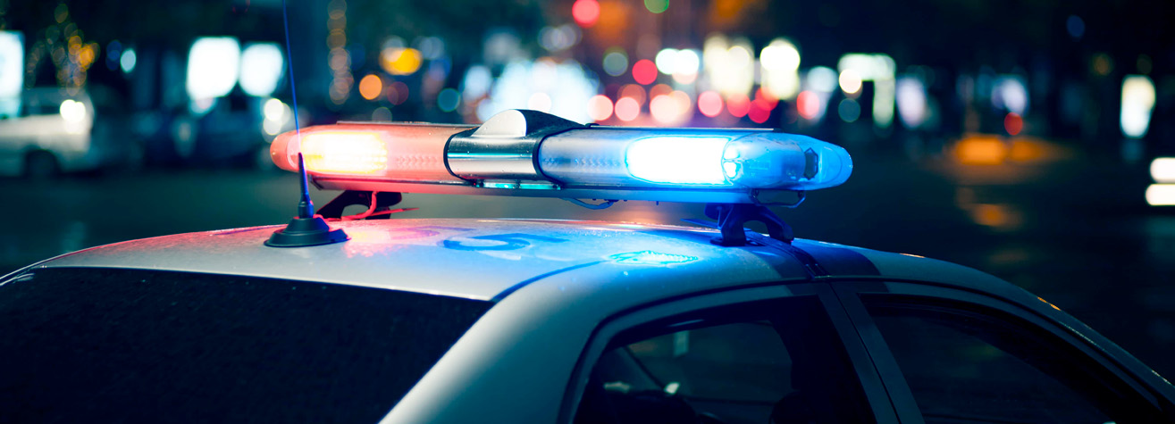 A police car with flashing red and blue lights is parked on a city street at night. The background is blurred with bokeh lights from streetlamps and buildings, creating a vibrant urban atmosphere.