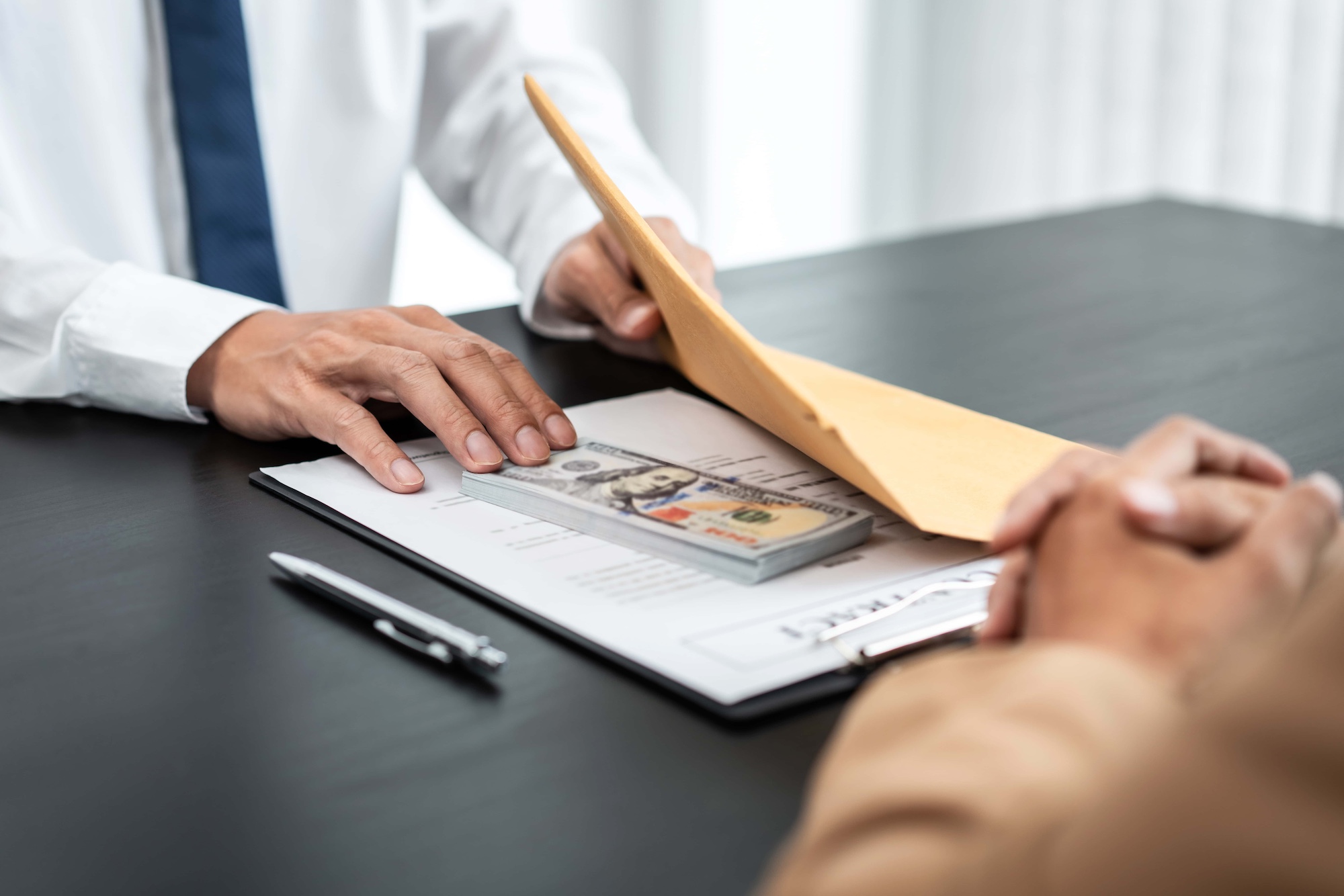 Two individuals sit at a table. One hands over an envelope with paperwork and a stack of U.S. hundred-dollar bills. A pen is placed on the table beside the documents.