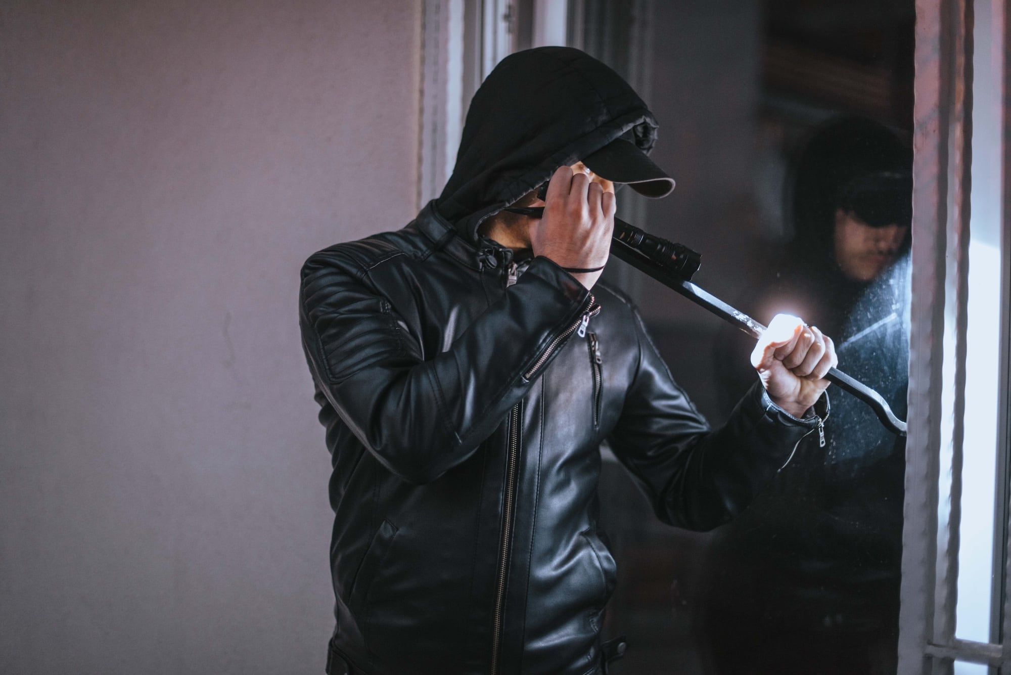 A person in a hooded jacket and cap stands in front of a glass door at night, holding a flashlight and a crowbar. The scene suggests a potential break-in.
